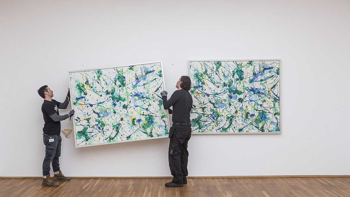 © Image: David von Becker Two people are hanging a painting on a wall next to a painting that already is hung up. 