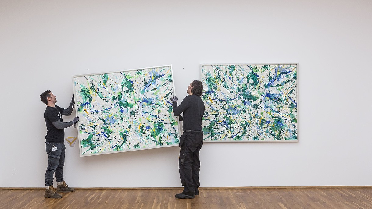 © Image: David von Becker Two people are hanging a painting on a wall next to a painting that already is hung up. 