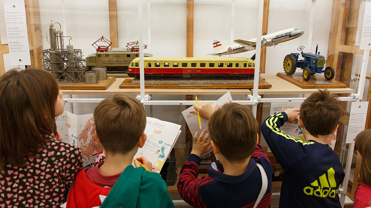  Children are standing in front of a display showcasing different forms of transportation. They hold printed maps on which they are writing.