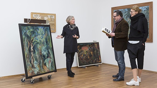 A woman is showing two people a painting that is standing on the ground. The man is reading about the painting in a book.
