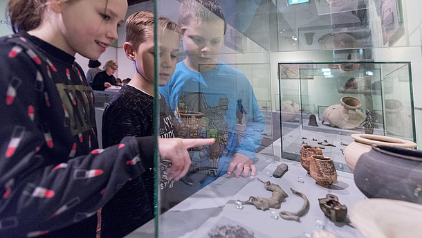 Three kids look at objects placed in a monter. The girl points at an object and explains it to the two boys. 