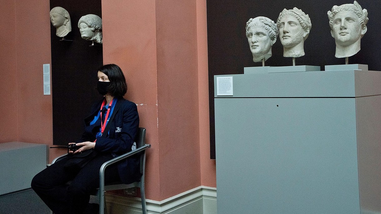 © Majority World CIC / Alamy Stock Foto A security officer, wearing a face mask, sits next to ancient stone busts inside a museum space