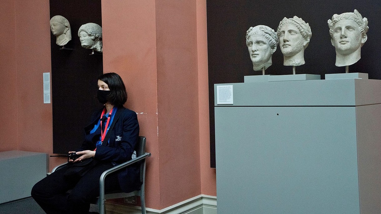 © Majority World CIC / Alamy Stock Foto A security officer, wearing a face mask, sits next to ancient stone busts inside a museum space